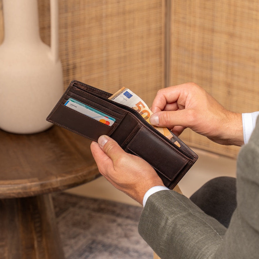 Man holding a brown wallet with cards against a wooden table and woven wall background