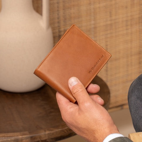 Man holding a cognac leather wallet next to a white vase on a wooden table.