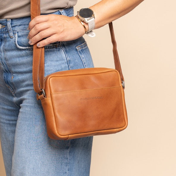 Woman holding a cognac leather crossbody bag against a beige background