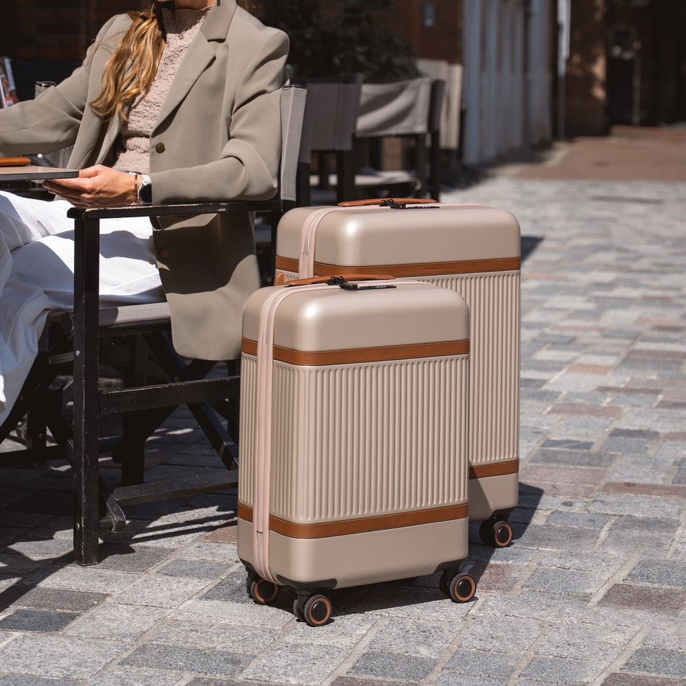 Two beige suitcases with brown accents on a stone pavement, with a person sitting at a table in the background.