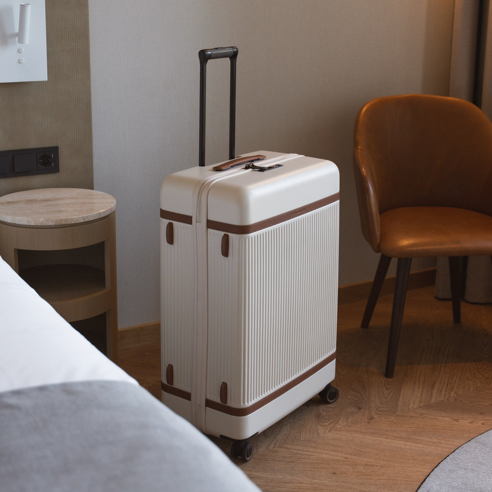 Off white suitcase with brown accents on a wooden floor in a room with a chair and bed.