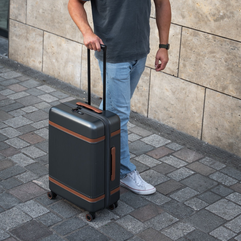 Person holding a grey suitcase with brown accents on a sidewalk.