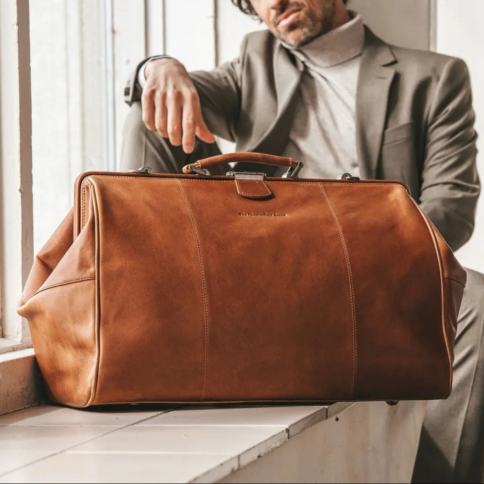 Man in gray suit holding a cognac leather duffel bag by a window.