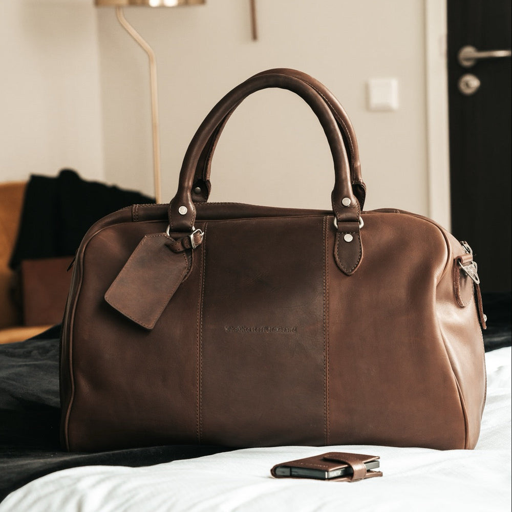 Brown leather duffel bag with tag on a bed in a room.