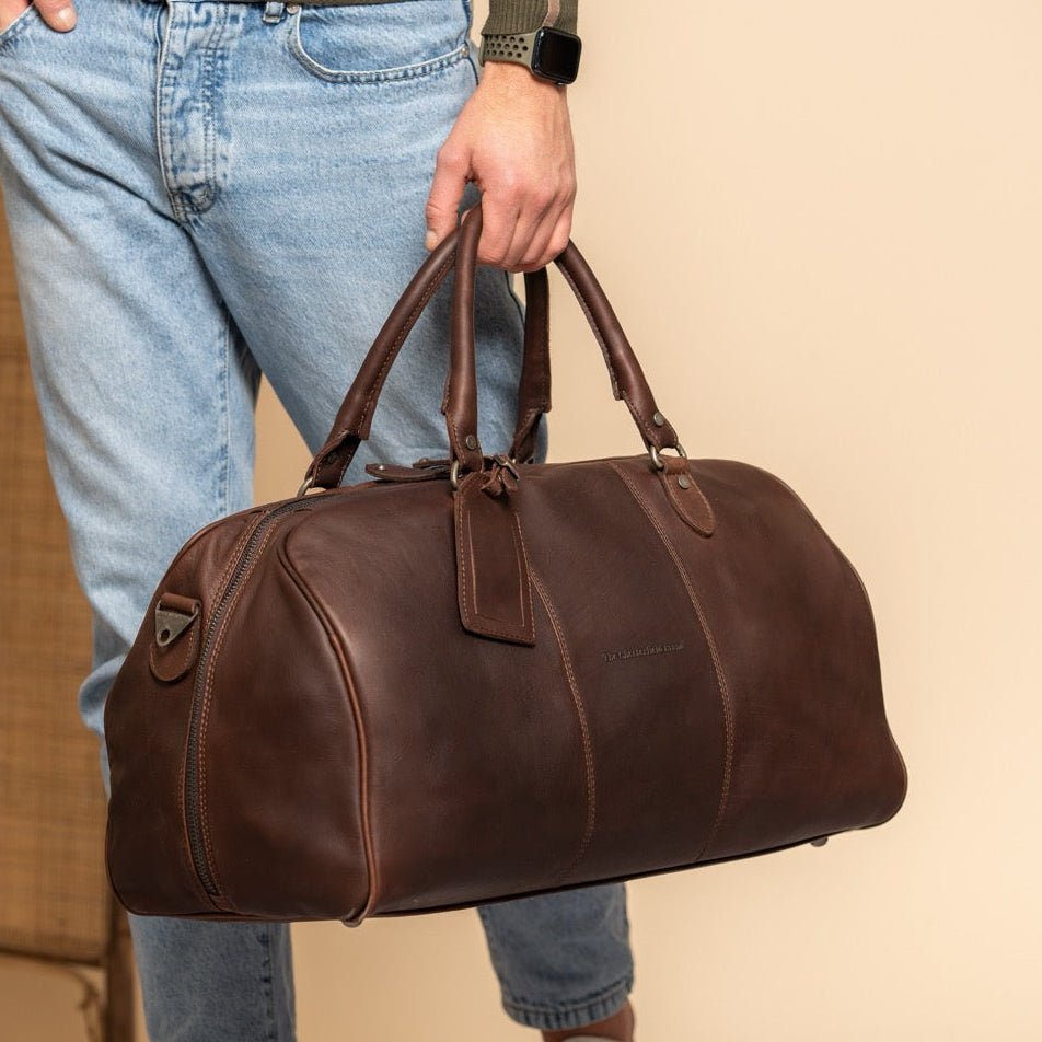 Person holding a brown leather duffel bag against a neutral background.
